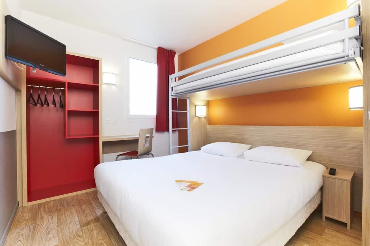A hotel room with a bunk bed, a desk with a chair, a TV mounted on the wall, and a bedside table with a lamp.