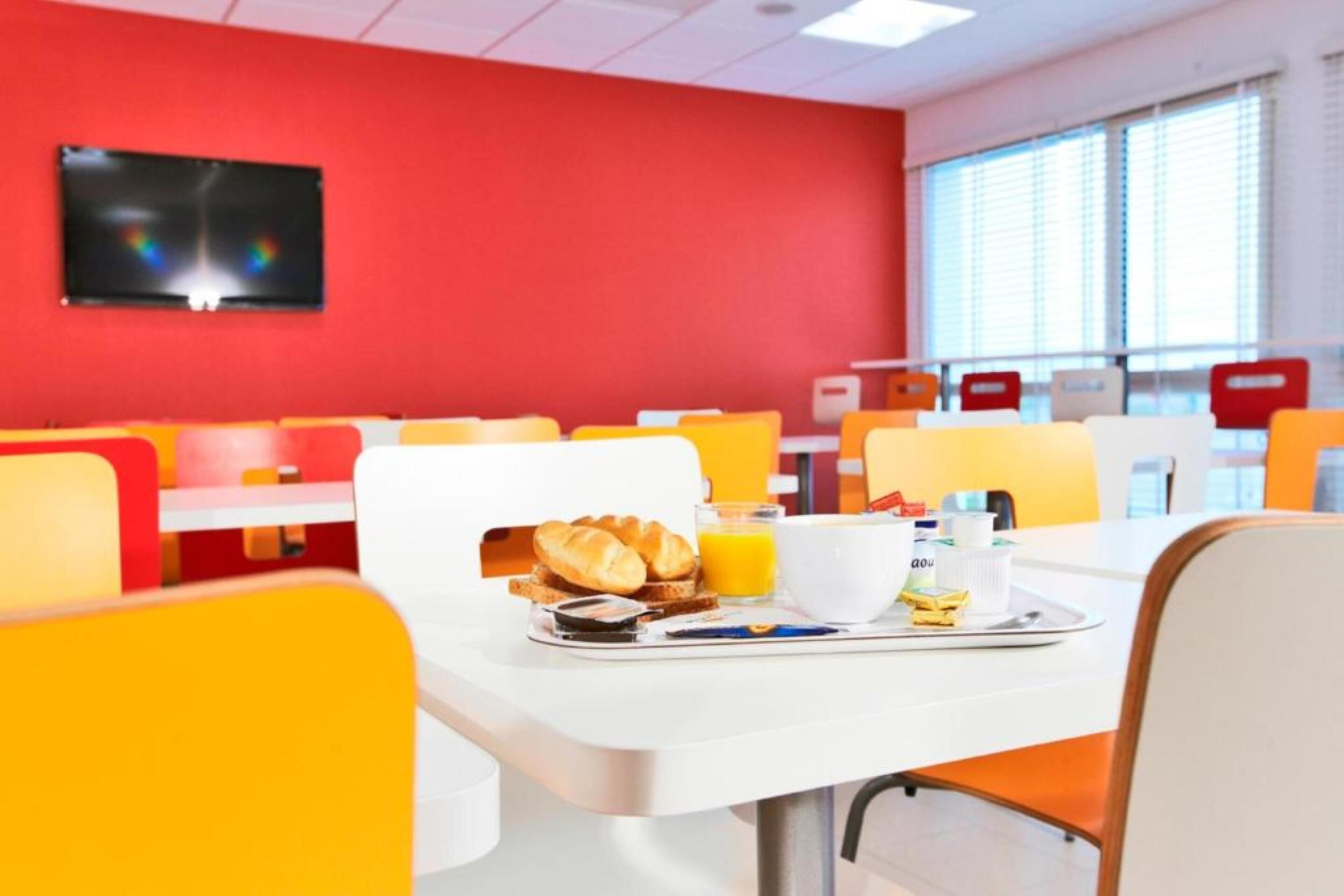 A brightly colored dining area with orange and red chairs, a white table with breakfast items, and a TV mounted on the red wall.