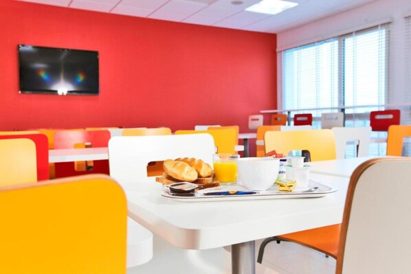 A brightly colored dining area with orange and red chairs, a white table with breakfast items, and a TV mounted on the red wall. - Hôtel Première Classe Caen Nord - Mémorial (Saint-Contest)