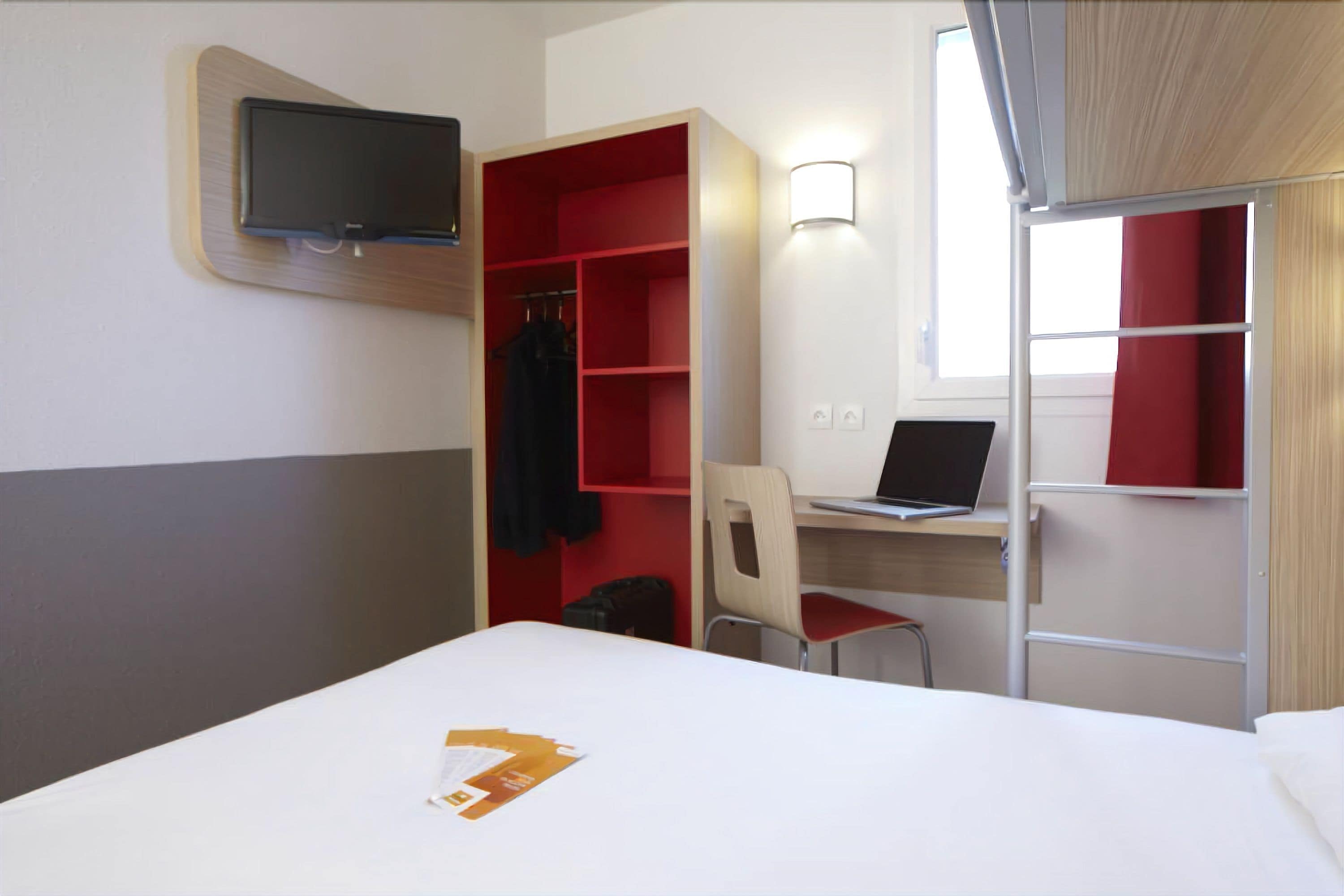 Een hotelkamer met een bed, een bureau met een laptop, een stoel, een aan de muur bevestigde televisie en een kast met rode planken.