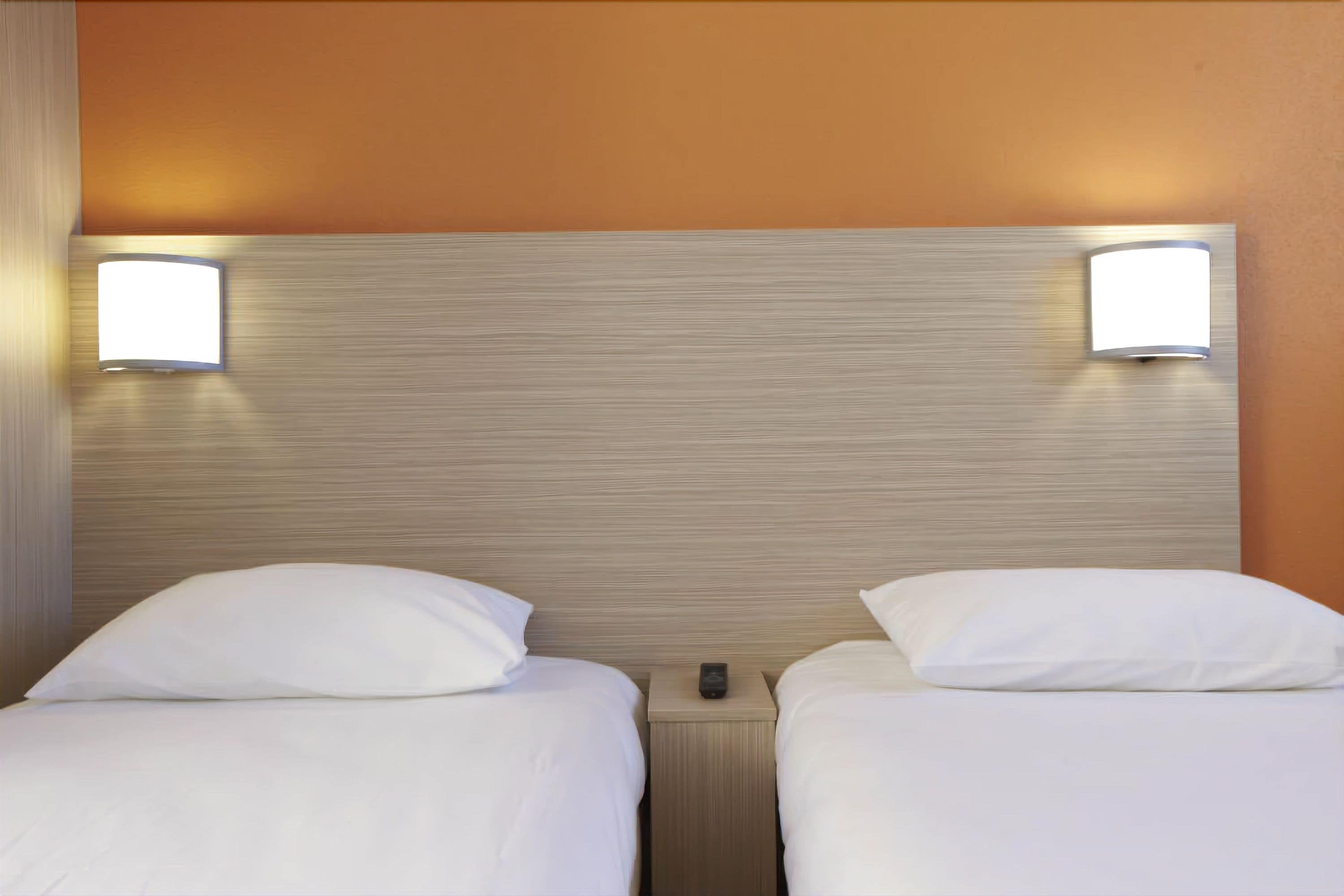 A hotel room with two single beds, each with a white pillow and a nightstand with a remote control.