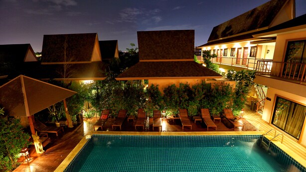 A swimming pool with lounge chairs and umbrellas, surrounded by greenery and buildings with sloped roofs. - Ploykhumthong Boutique Resort (Bangkok)