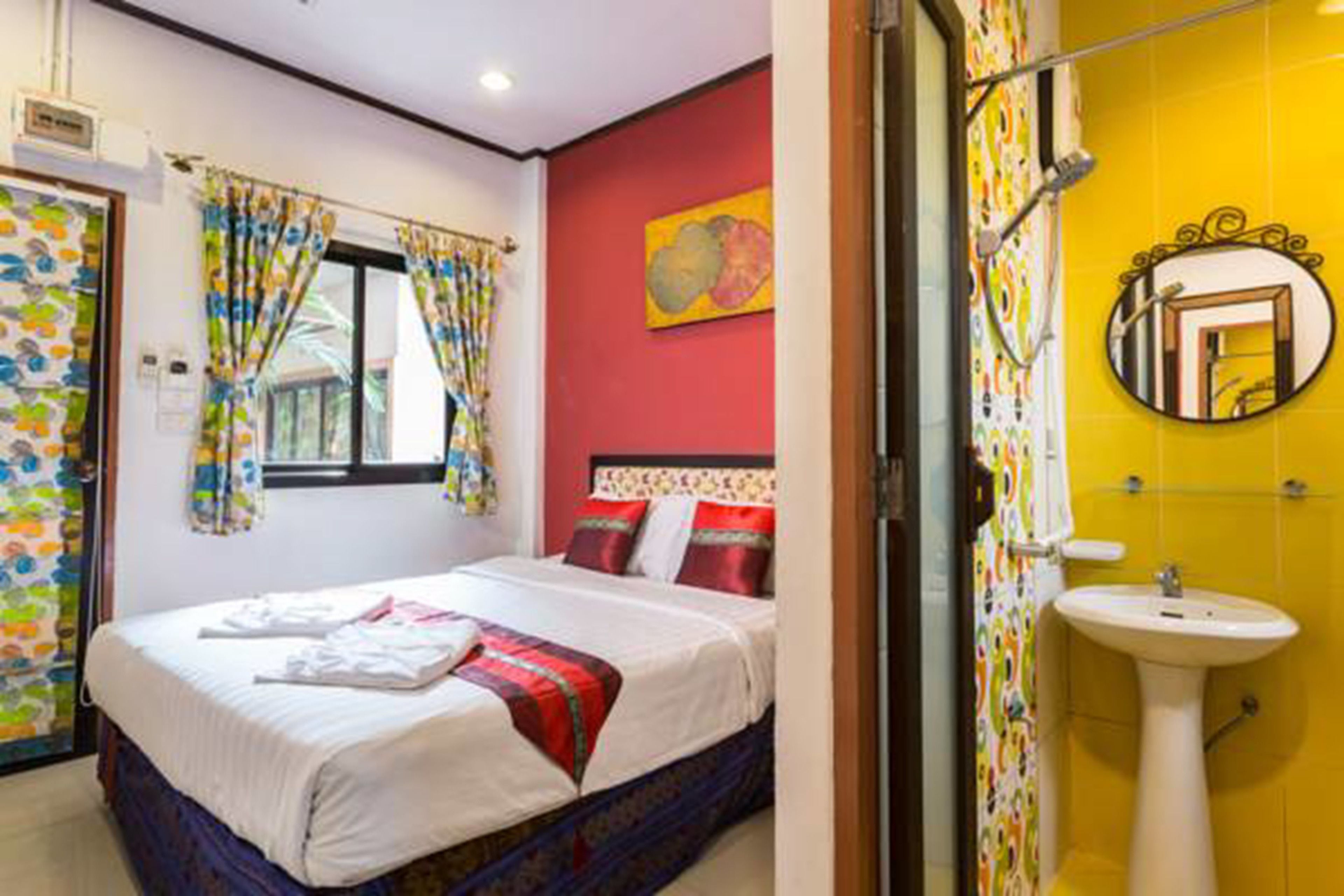 A hotel room with a bed, a window with curtains, a red wall, and a yellow bathroom with a sink and mirror.