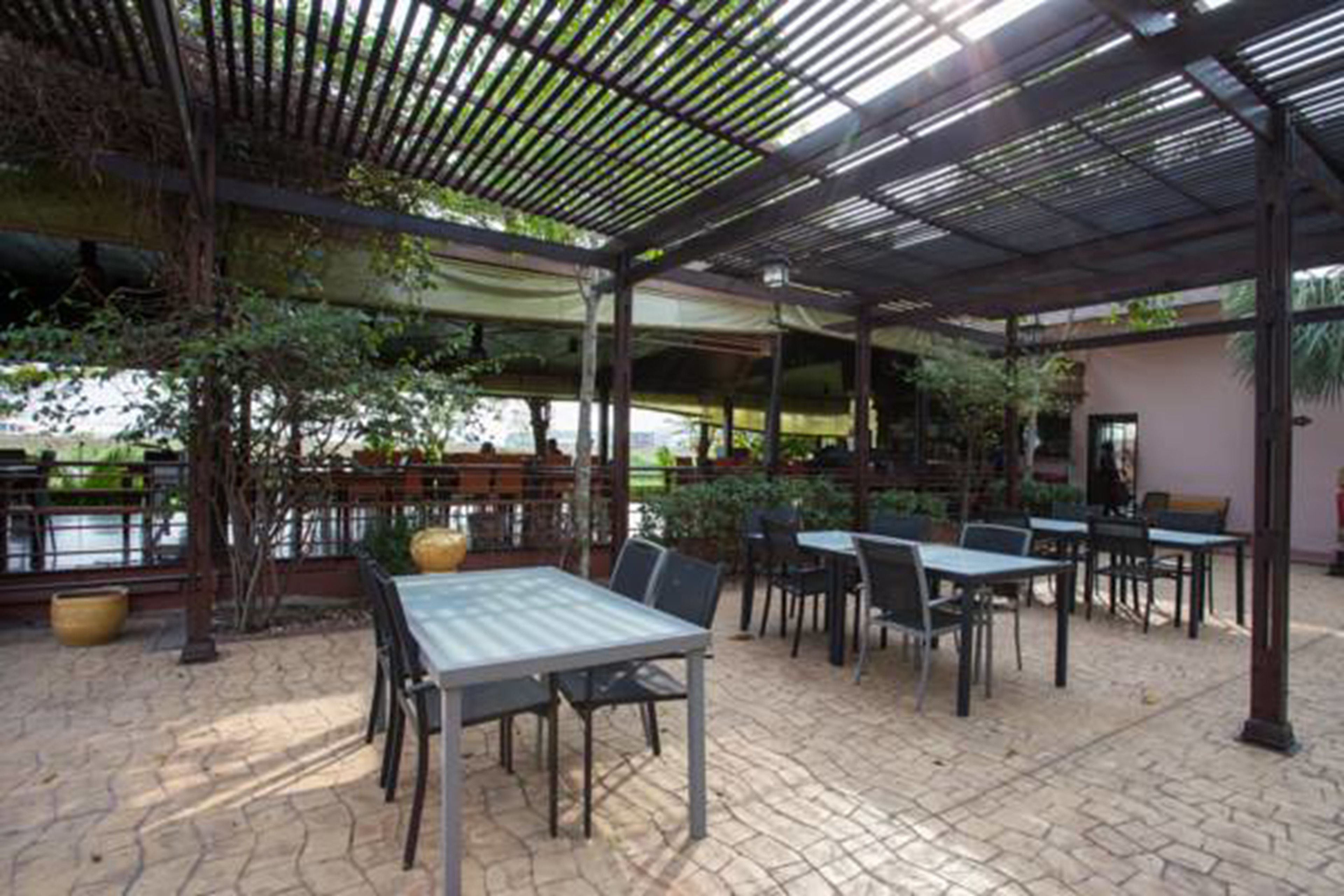 Un espace extérieur aménagé en terrasse, avec des tables et des chaises, recouvert d’un toit en métal, entouré de verdure et offrant une vue sur une étendue d’eau.
