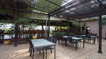 Un espace extérieur aménagé en terrasse, avec des tables et des chaises, recouvert d’un toit en métal, entouré de verdure et offrant une vue sur une étendue d’eau.