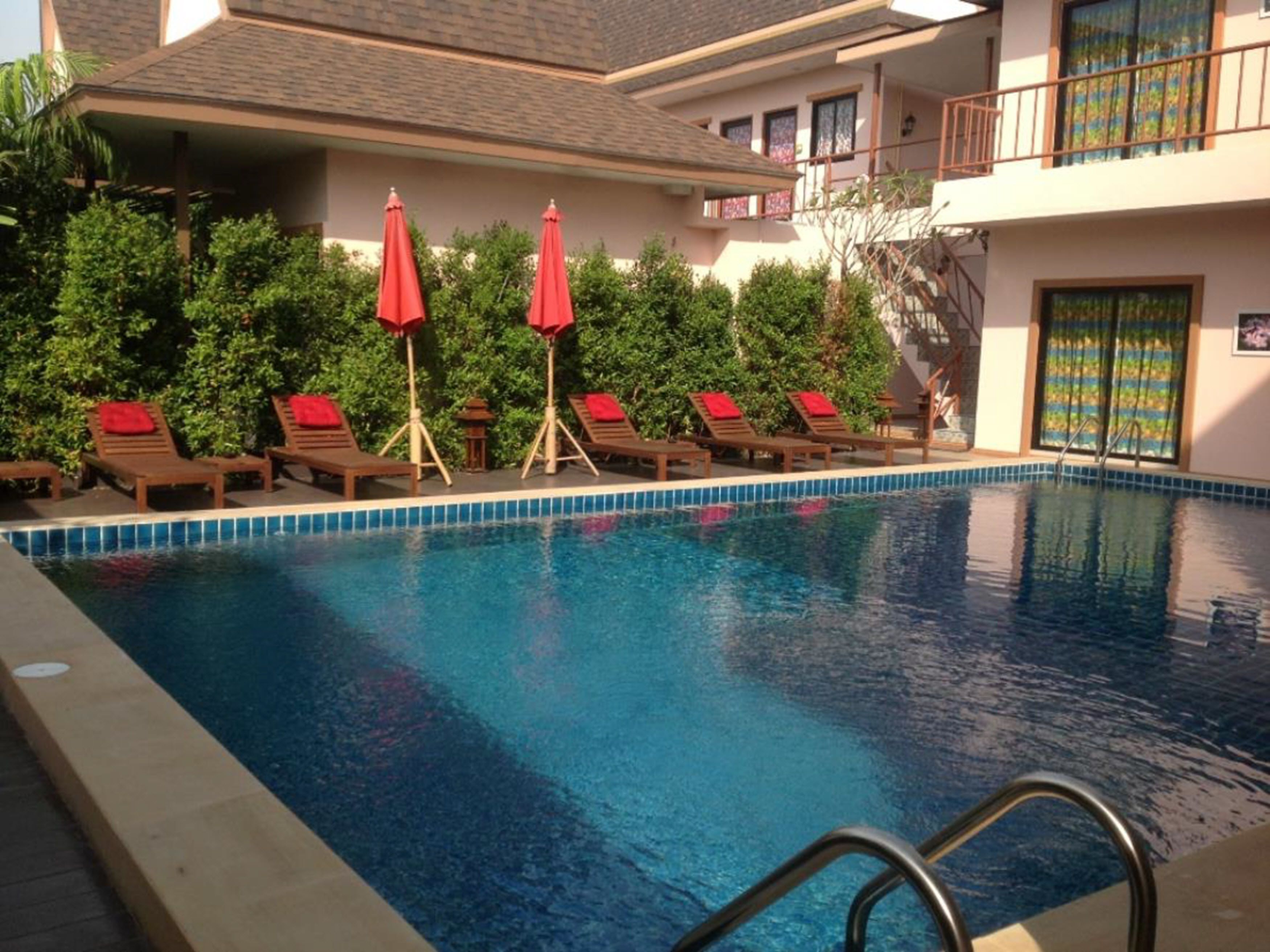 A swimming pool with lounge chairs and red umbrellas, surrounded by greenery and a building with a balcony.
