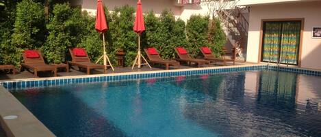 A swimming pool with lounge chairs and red umbrellas, surrounded by greenery and a building with a balcony.