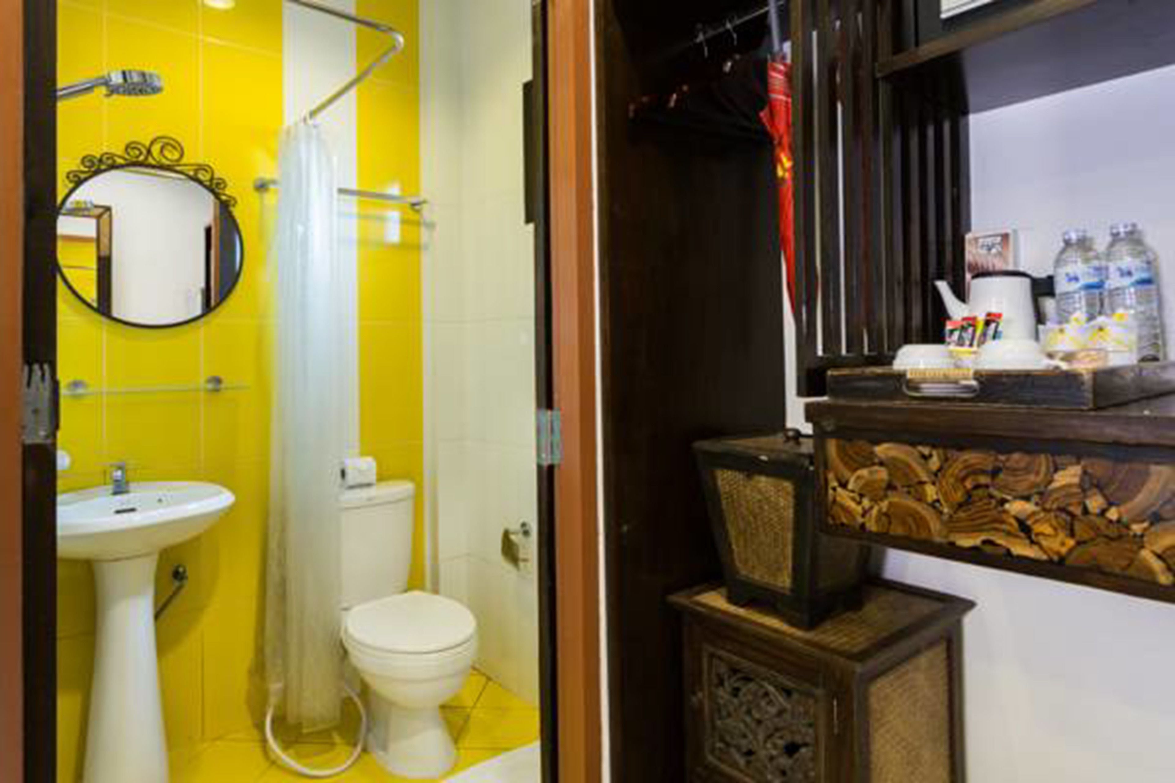 A bathroom with a yellow tiled wall, a white sink, a toilet, and a mirror. There is a wooden shelf with toiletries.