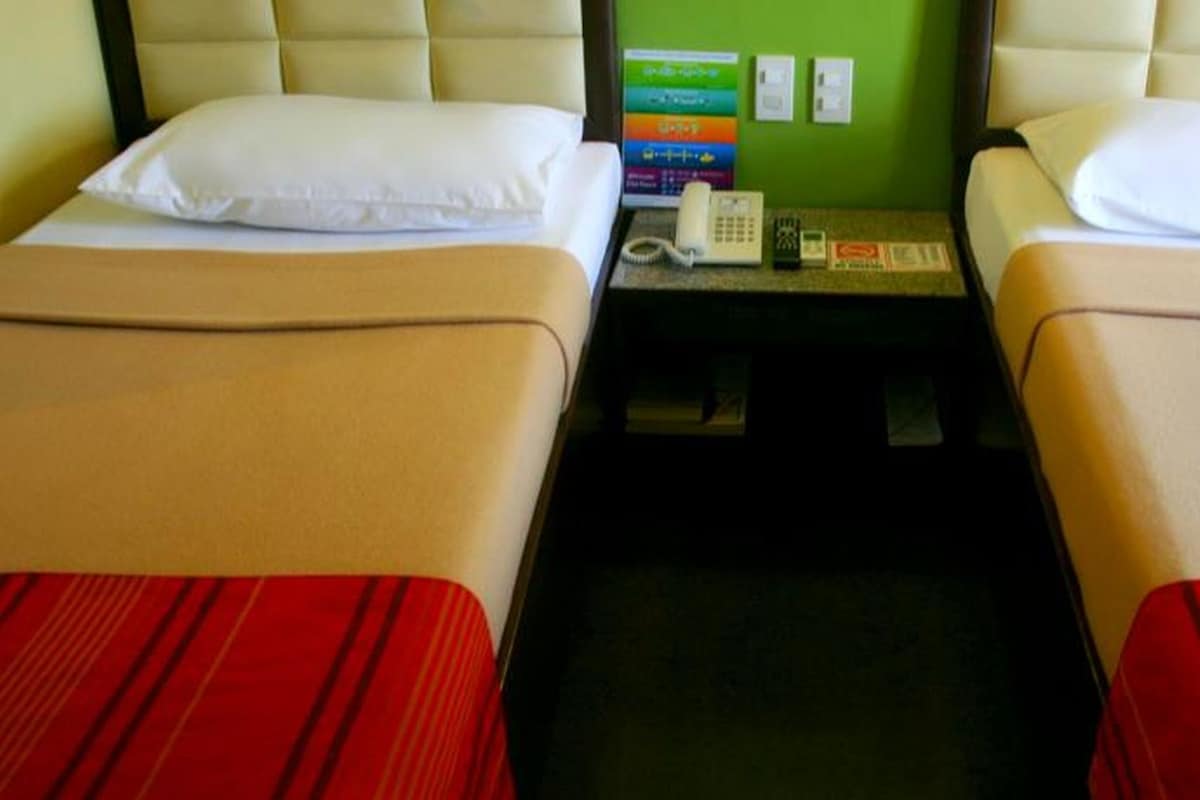 A hotel room with two beds, each with a pillow and a red-striped bedspread. There is a small table between the beds with a telephone and a wall-mounted light switch.