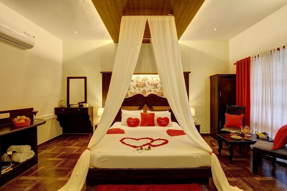 Un dormitorio con cama de dos columnas, cabecera de madera, mesita de noche con lámpara, una silla roja y una pequeña mesa con tetera y tazas.