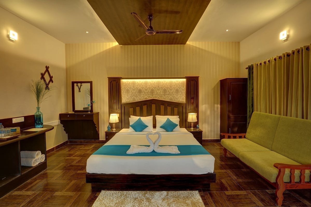 Habitación de hotel con una cama grande, cabecera de madera, dos mesitas de noche con lámparas, un sofá verde y suelo de madera.