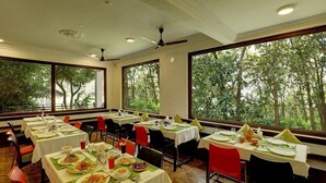 A restaurant with multiple tables, each set with plates, glasses, and cutlery. Large windows provide a view of trees outside. - Periyar Nest Resorts (Peermade)