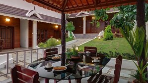 A covered outdoor seating area with a glass table, chairs, and potted plants. - Periyar Nest Resorts (Peermade)
