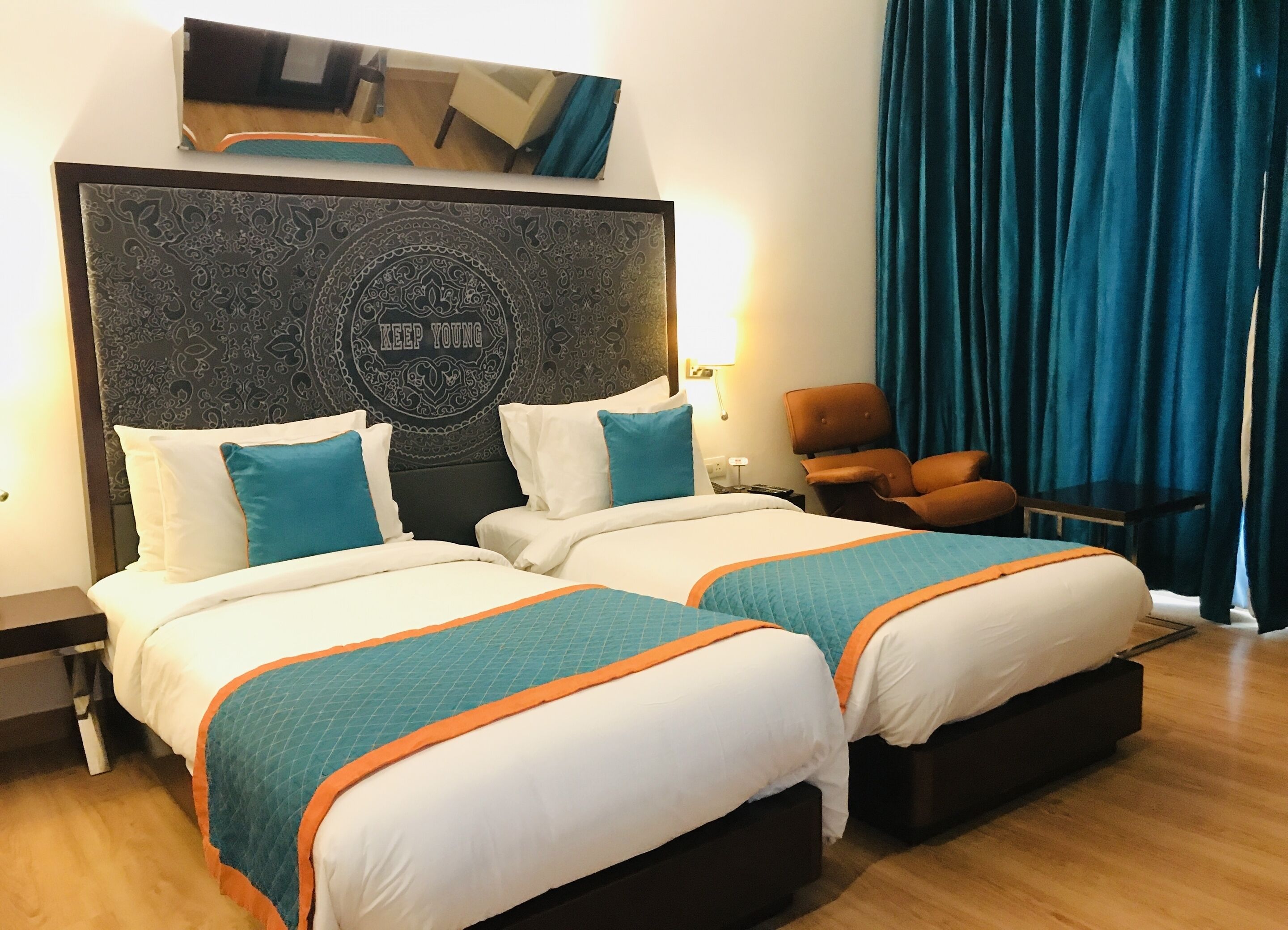 Habitación de hotel con dos camas, un cabecero grande con un diseño decorativo, una silla y un ventanal con cortinas.