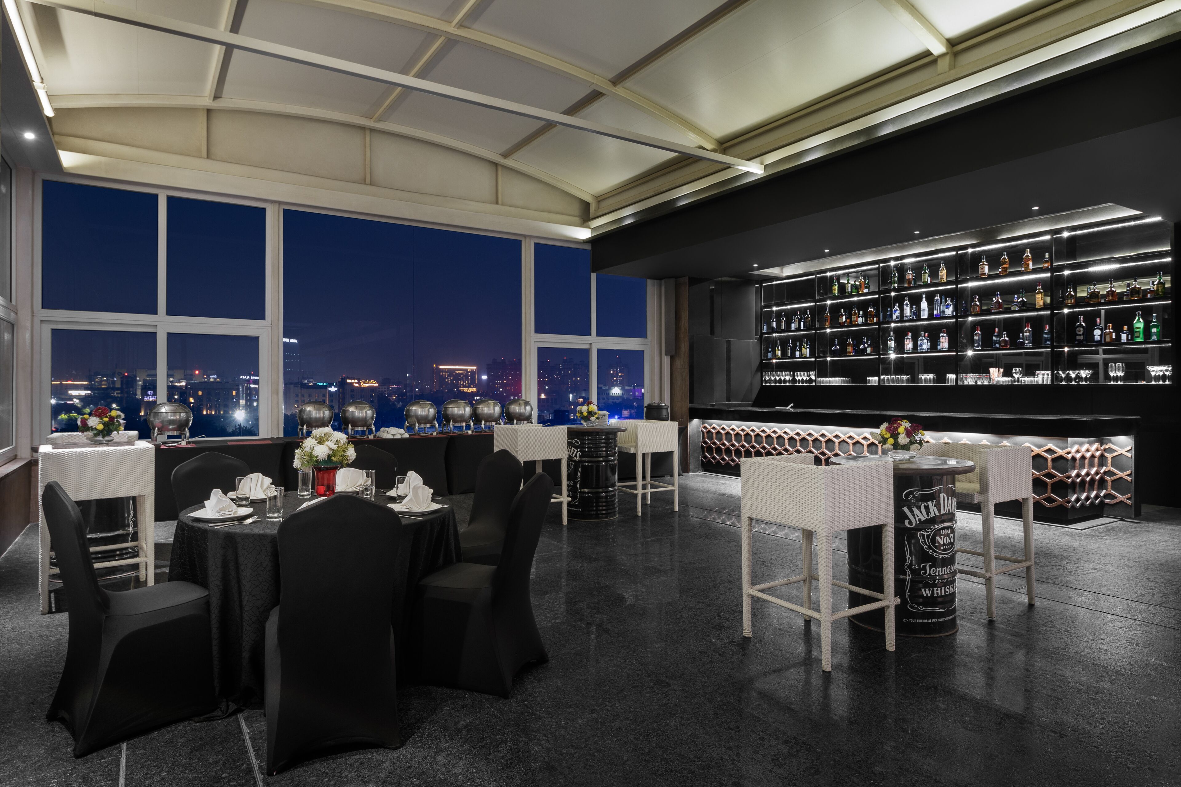 A modern bar area with a city view, featuring a well-stocked liquor shelf and a seating area with tables and chairs.