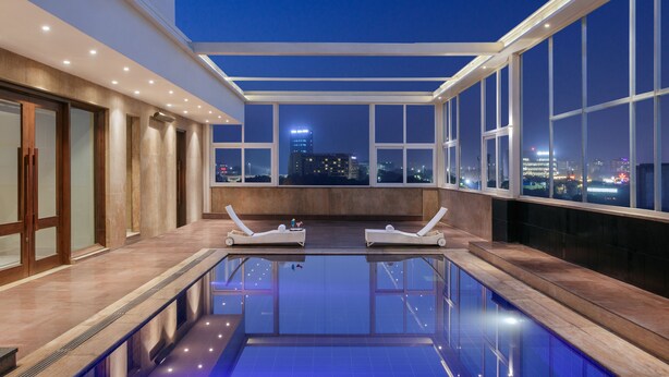 A rooftop pool area with two sun loungers, a glass barrier, and a city view at dusk. - Golden Tulip Gurgaon - Sector 29 (Gurugram)