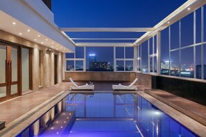 A rooftop pool area with two sun loungers, a glass barrier, and a city view at dusk. - Golden Tulip Gurgaon - Sector 29 (Gurugram)
