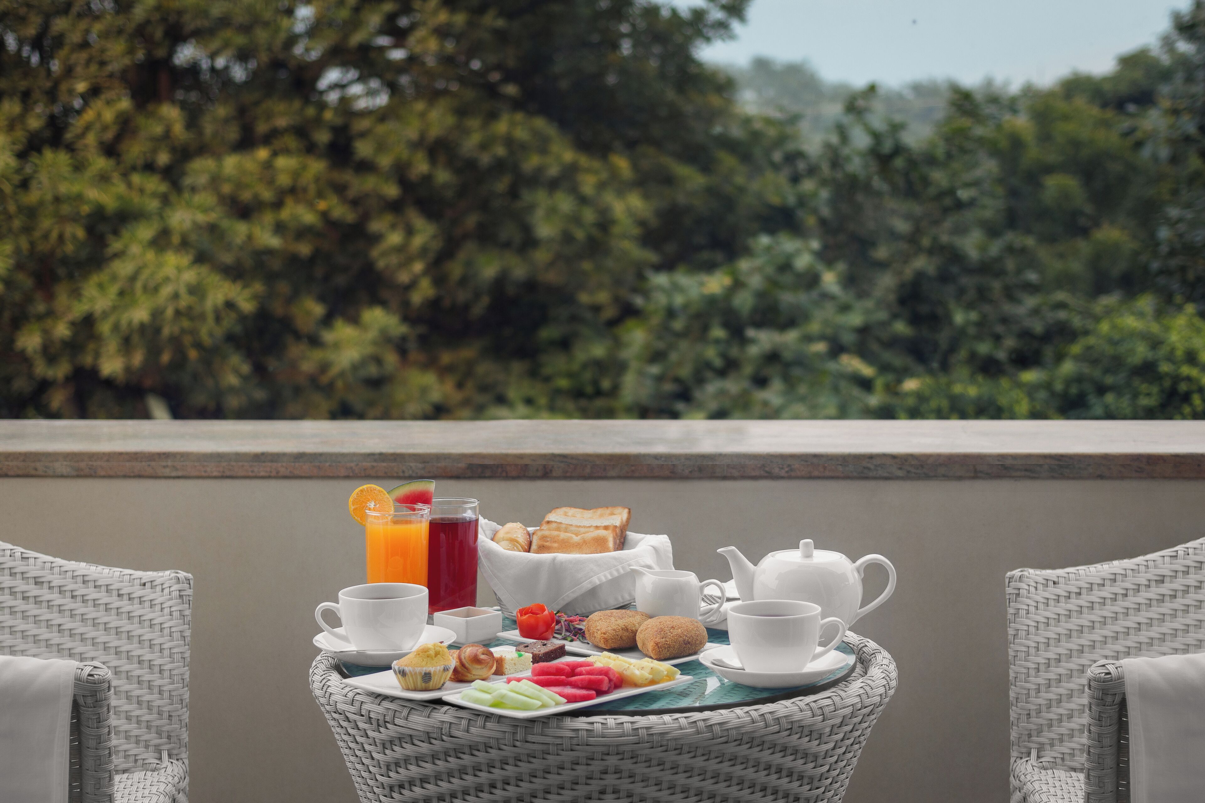Una mesa de mimbre con alimentos para el desayuno, incluyendo tostadas, fruta e infusiones, sobre un balcón con vistas a árboles.