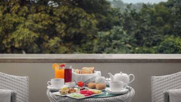 Una mesa de mimbre con alimentos para el desayuno, incluyendo tostadas, fruta e infusiones, sobre un balcón con vistas a árboles.