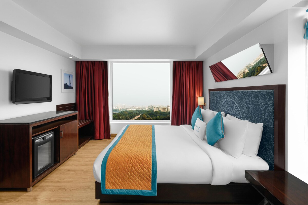 Habitación de hotel con cama, televisión, escritorio, ventana con vista a la ciudad y cabecero con diseño estampado.