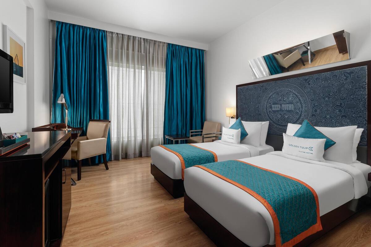 Habitación de hotel con dos camas, un escritorio, una silla, un televisor y un ventanal grande con cortinas.
