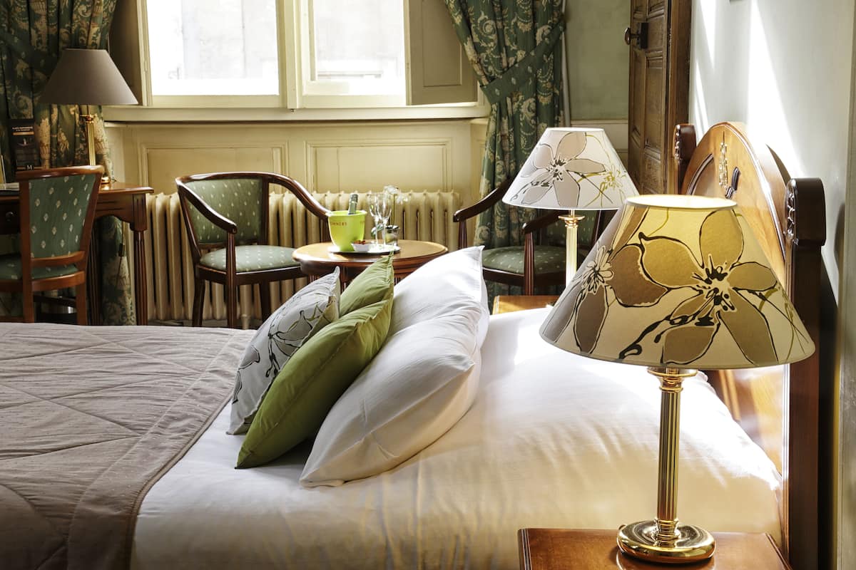 A neatly made bed with pillows, a bedside lamp, and a view of a dining area with chairs and a table.