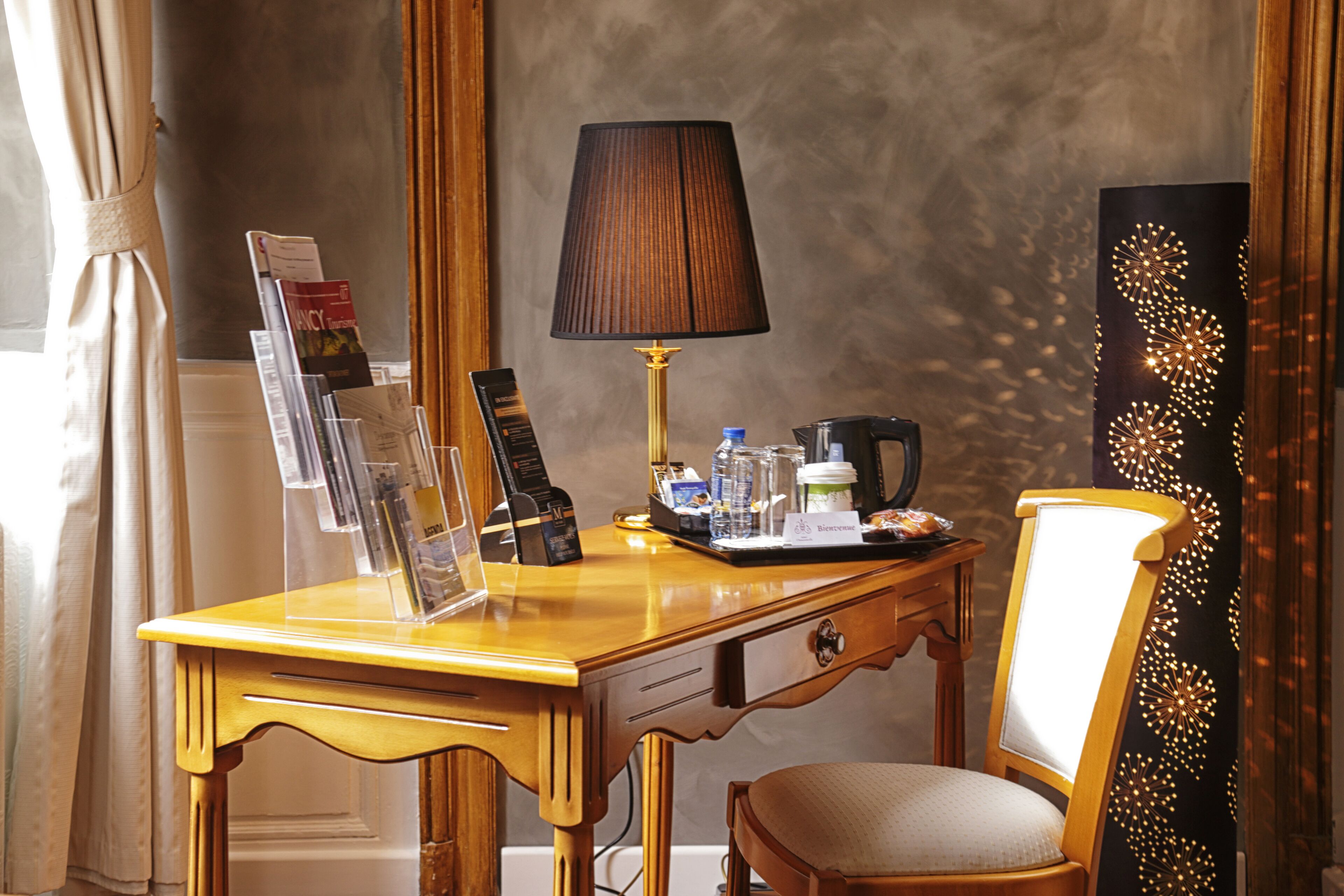 A hotel desk with a lamp, a tray with a coffee pot, a water bottle, a glass, and a magazine rack.