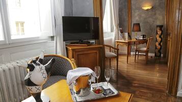 A hotel room with a wooden dining table, a television, a decorative sculpture, and a tray with a bottle, glasses, and fruit.
