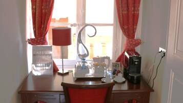 A small office with a desk, chair, lamp, coffee maker, and a window with curtains.