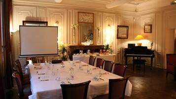 A formal dining room with a long table set for a meal, surrounded by chairs. There is a piano in the background and a whiteboard on the left.