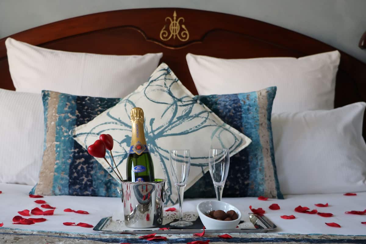 A bed with a wooden headboard, two white pillows, a decorative pillow with a blue pattern, a champagne bottle, two champagne flutes, a bowl of chocolates, and scattered rose petals.