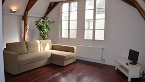 A cozy living room with a beige sectional sofa, a small white TV stand, and a large window with white shutters. - Rembrandtplein B&B (Amsterdam)