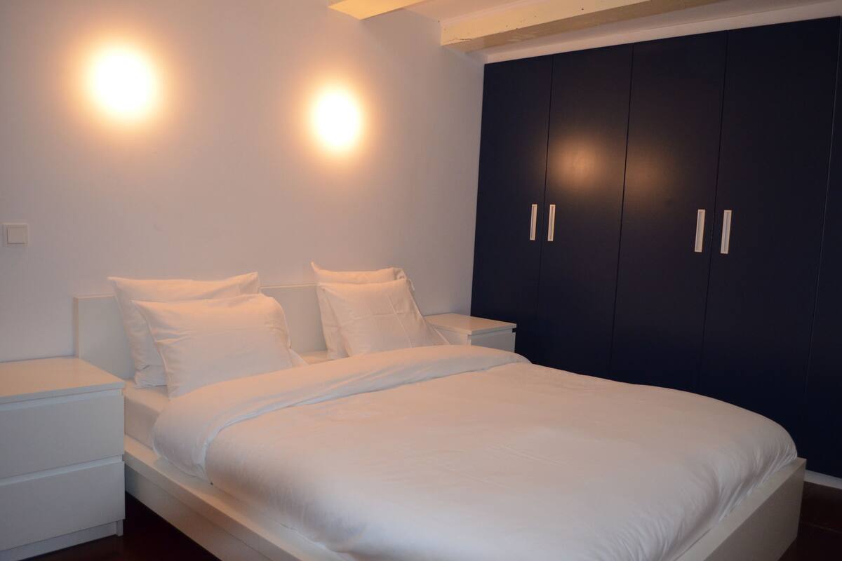A bedroom with a large bed, two bedside tables, and a dark wardrobe.