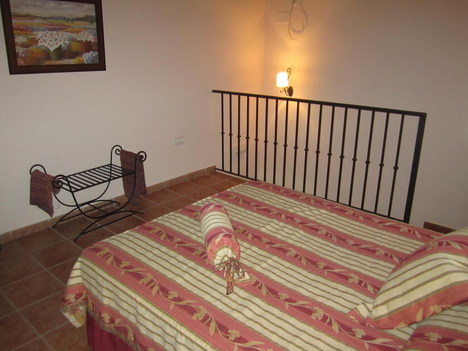 Un dormitorio con una cama, una lámpara de pared, un estante pequeño con una toalla y un cuadro en la pared.