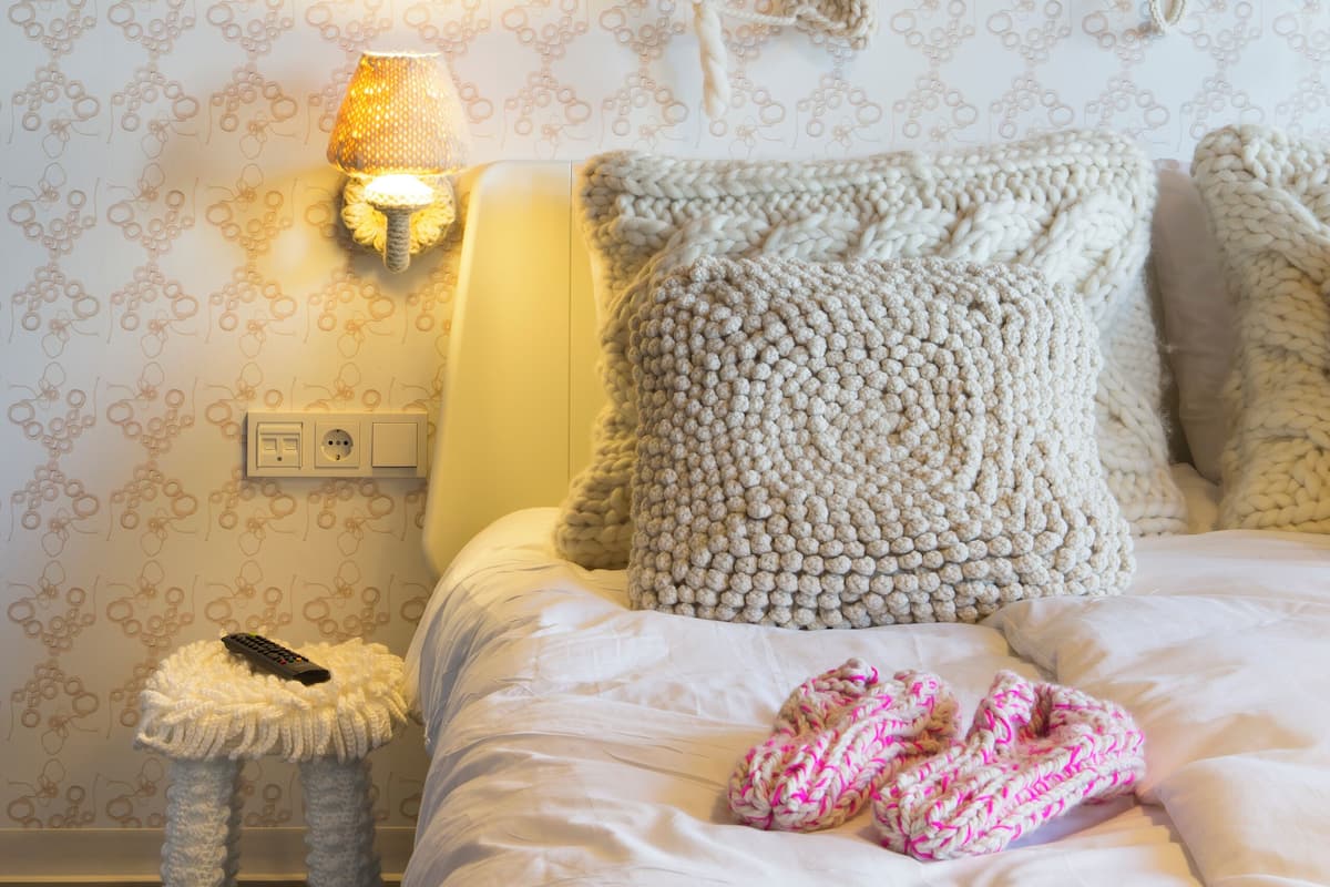 Un dormitorio acogedor con una cama, un taburete tejido y una lámpara de pared.
