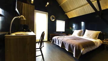 Un dormitorio con un escritorio de madera, una silla, una cama con manta, una mesita de noche y un ventanal.