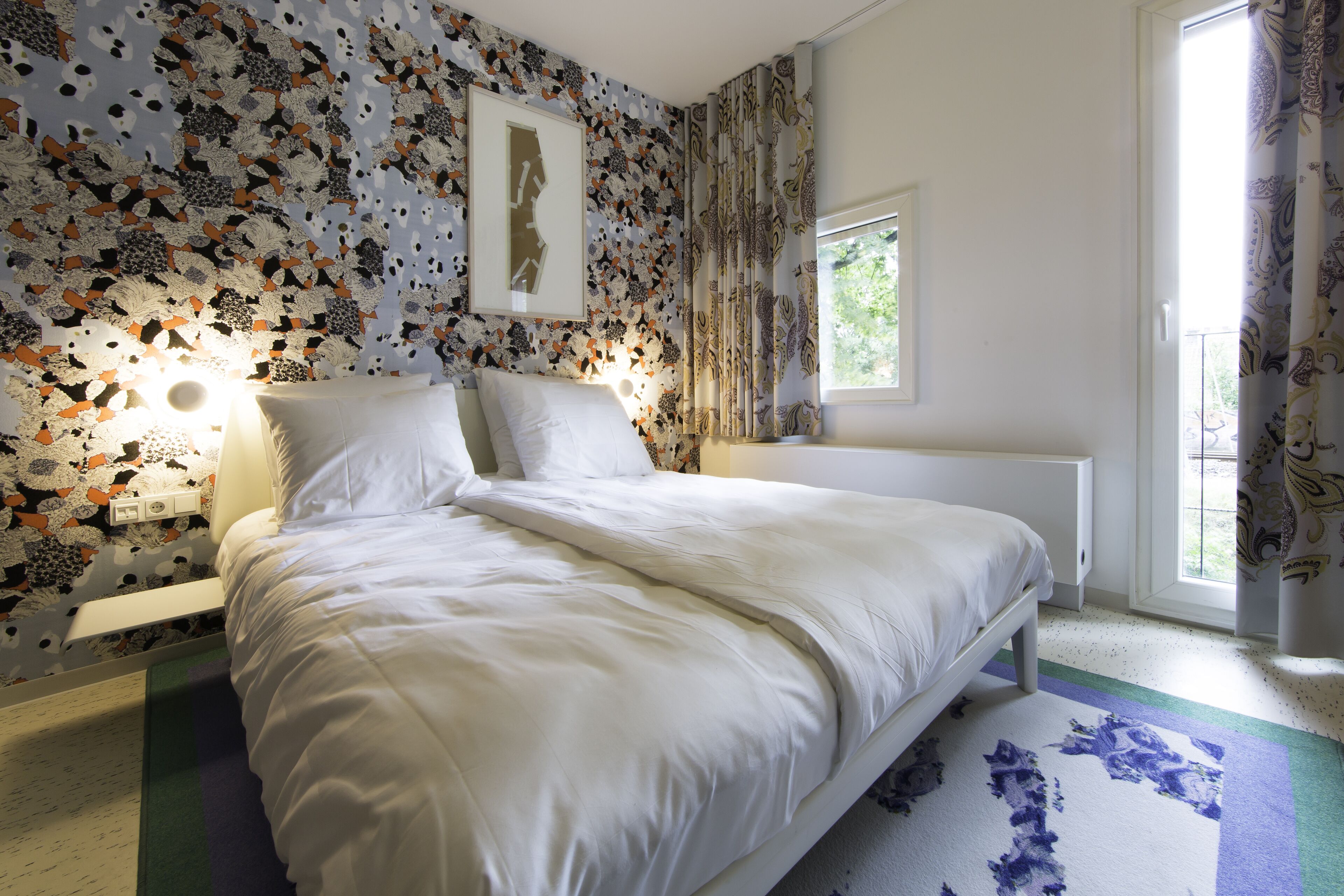 Un dormitorio con cama, ventana y pared con estampado floral.