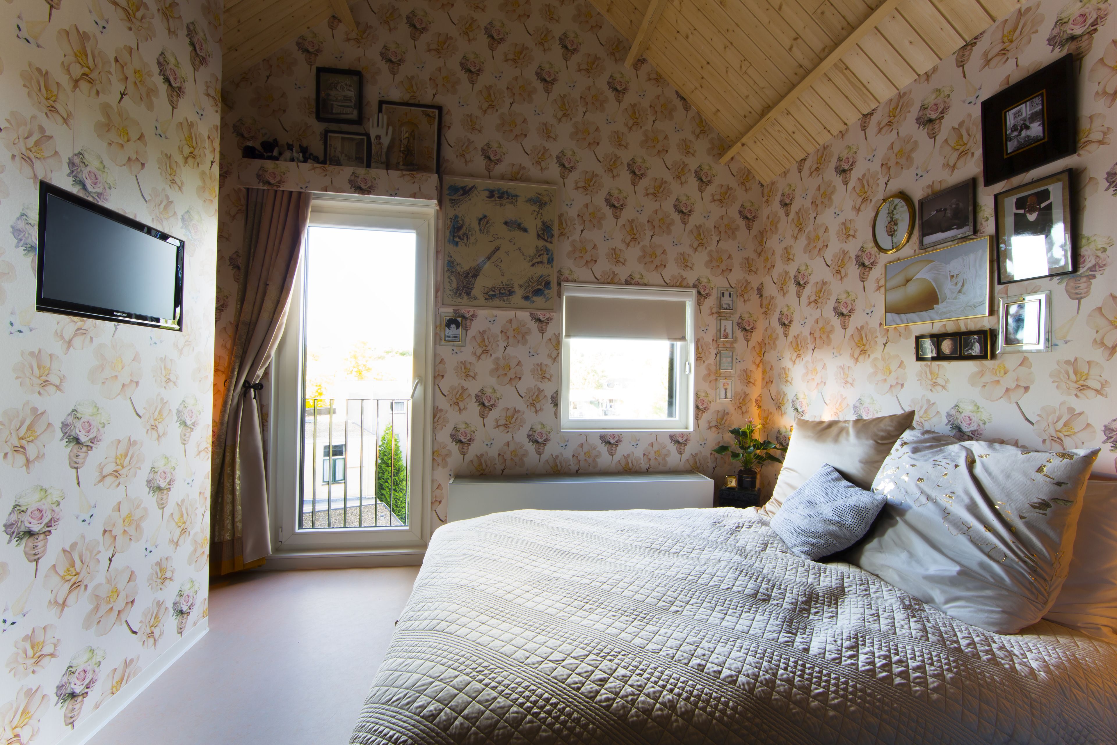 A bedroom with floral wallpaper, a bed with pillows, a TV, and a window with curtains.