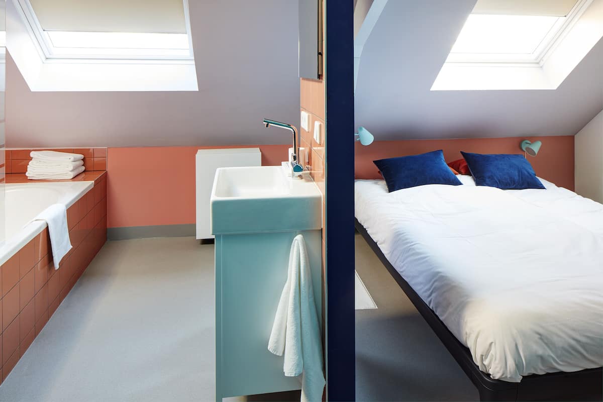 Un dormitorio moderno con tragaluz, cama con almohadas azules y rojas, lavamanos blanco y una pila de toallas blancas.