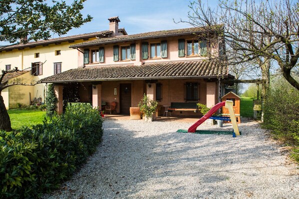 A two-story house with a gravel driveway, a wooden bench, and a red slide.