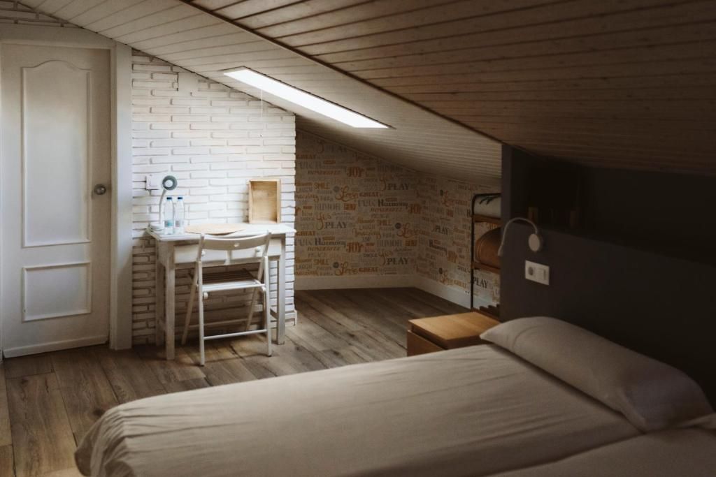 Un dormitorio con cama, escritorio y silla, una mesita pequeña y techo inclinado con tragaluz.