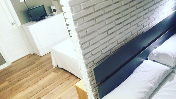 A room with a brick wall, a bed with white bedding, a wooden floor, a white dresser with a television on top, and a small table with a phone and a photo frame. - Hostal Le vintage (Ávila)