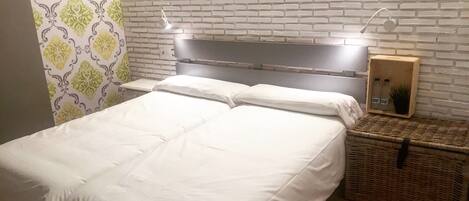 Una cama con ropa de cama blanca, cabecera con iluminación integrada, una pequeña cesta de mimbre y una pared con diseño de ladrillo.