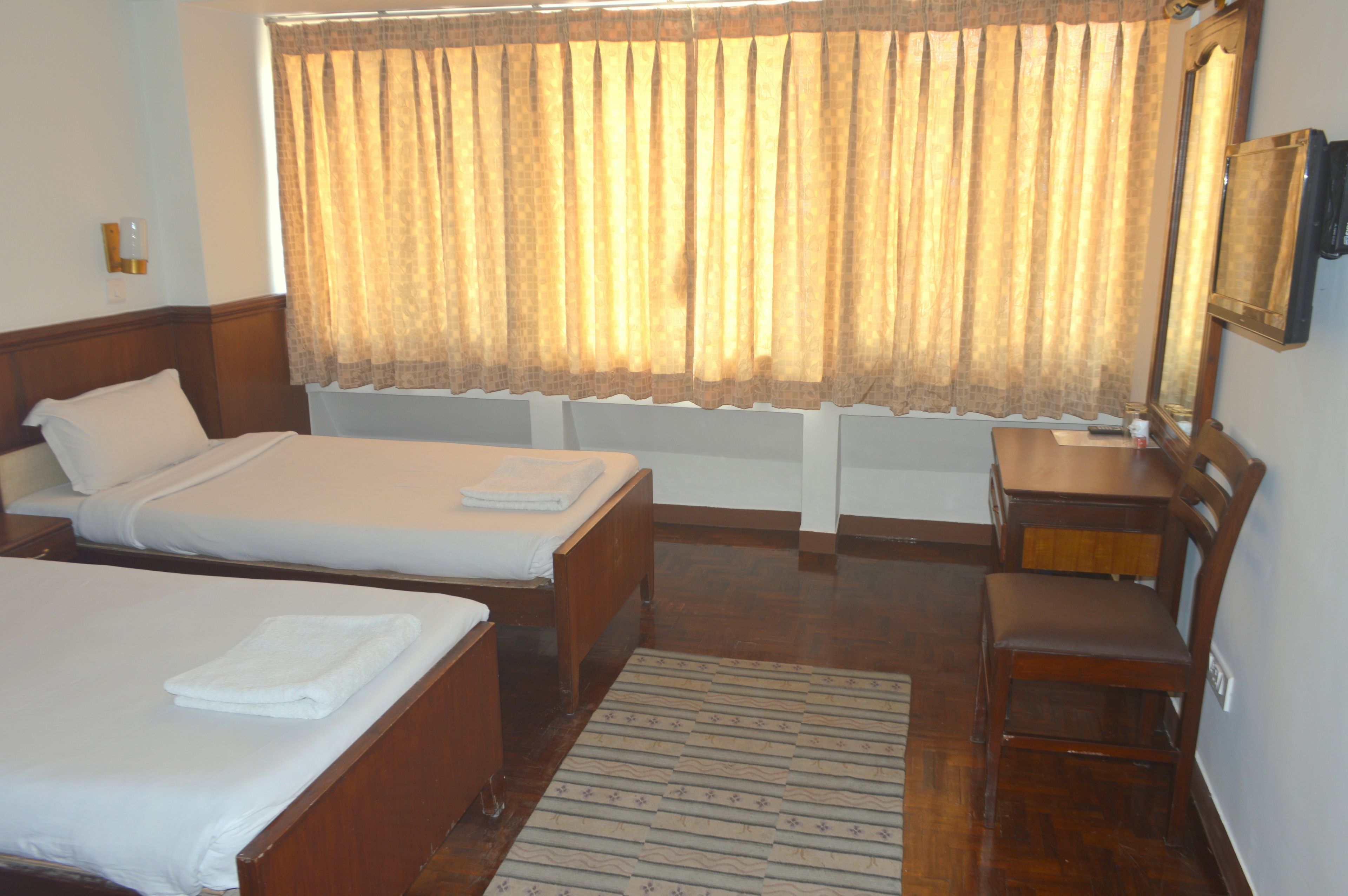 Habitación de hotel con dos camas individuales, un escritorio de madera, una silla, un espejo y una ventana con cortinas.