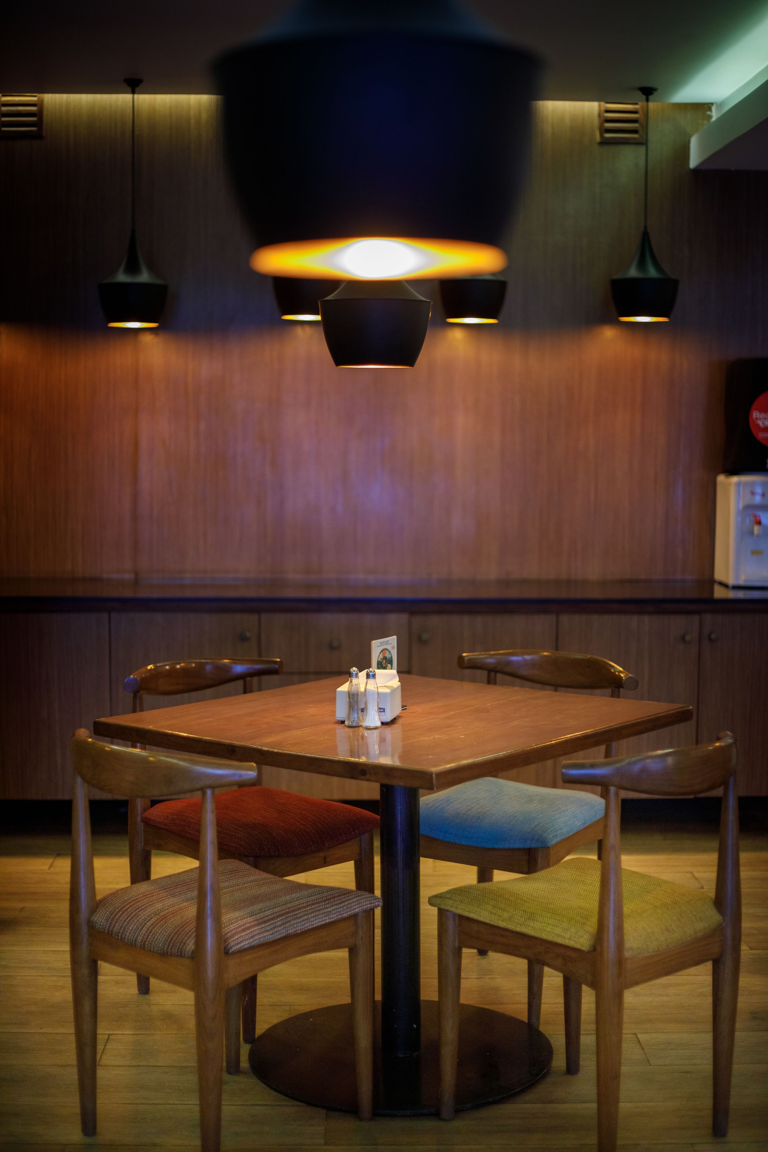 A dining area with a wooden table and chairs, pendant lighting, and a wooden wall.