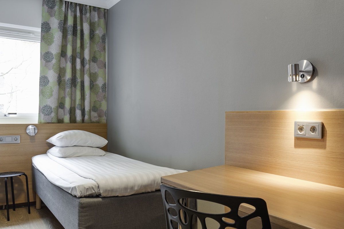 Habitación de hotel con cama, escritorio, silla, ventana con cortinas y un enchufe en la pared.