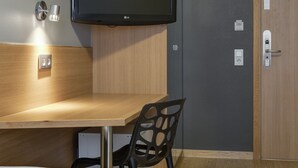 Habitación de hotel con cama, escritorio con silla, televisor de pantalla plana montado en la pared y puerta con picaporte.