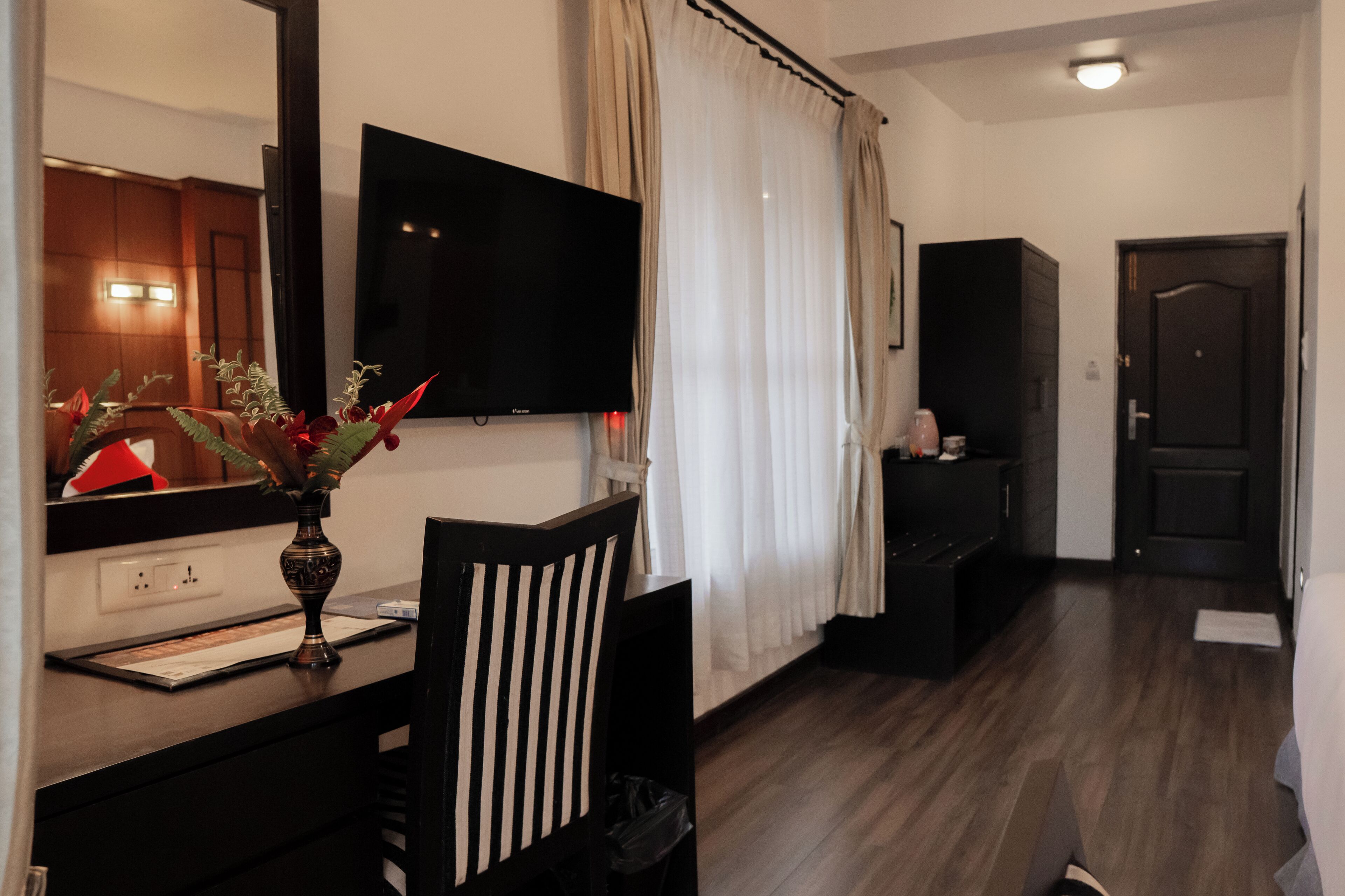 Une chambre d’hôtel équipée d’un téléviseur à écran plat fixé au mur, d’un bureau agrémenté d’un vase de fleurs, d’une chaise, d’une armoire et d’une porte.