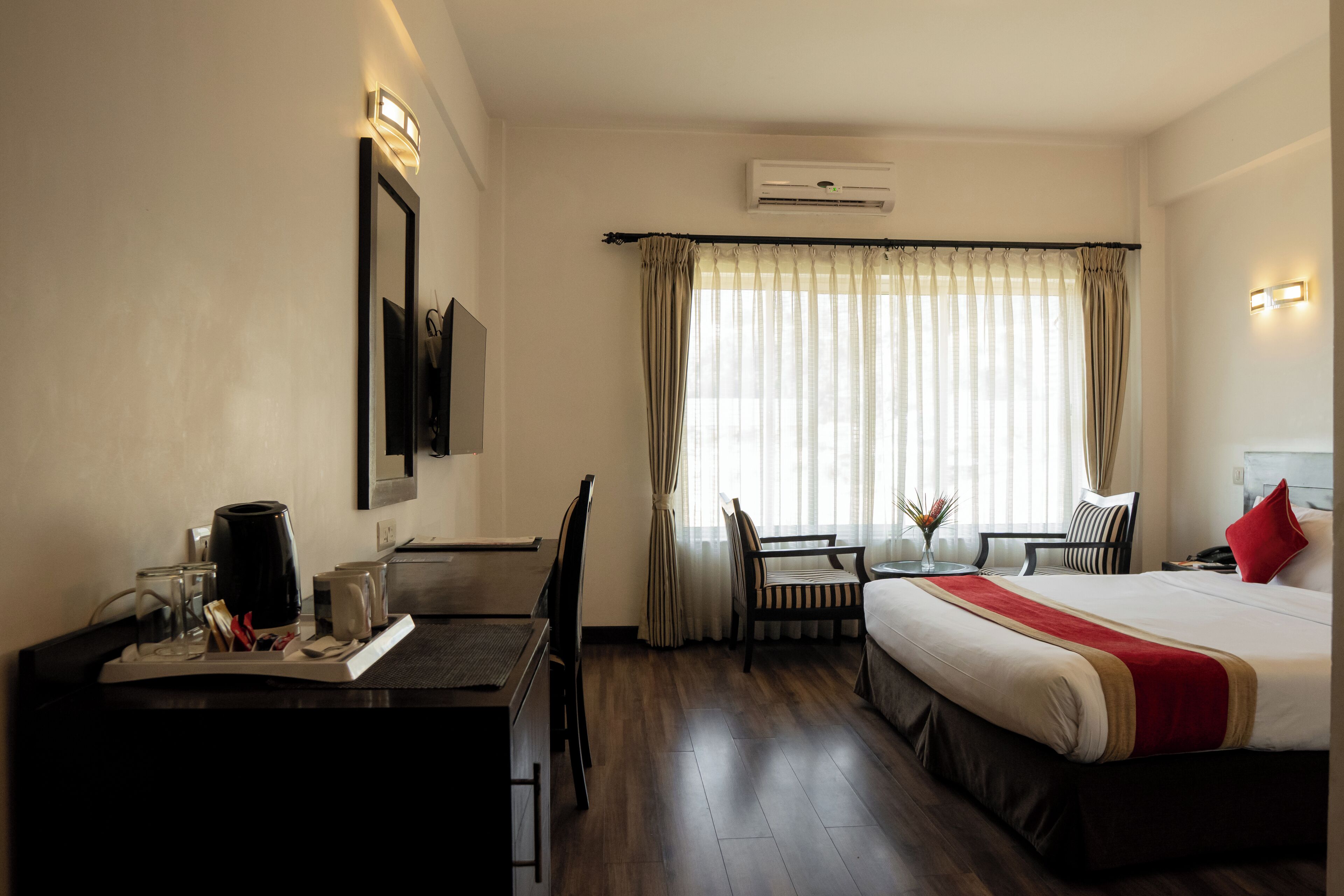 A hotel room with a bed, a desk with a chair, a small table with a vase, and a window with curtains.