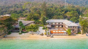 A beachfront property with a large building, a beach area with lounge chairs, and a forested hillside in the background. - Baan Ploy Sea (Rayong)